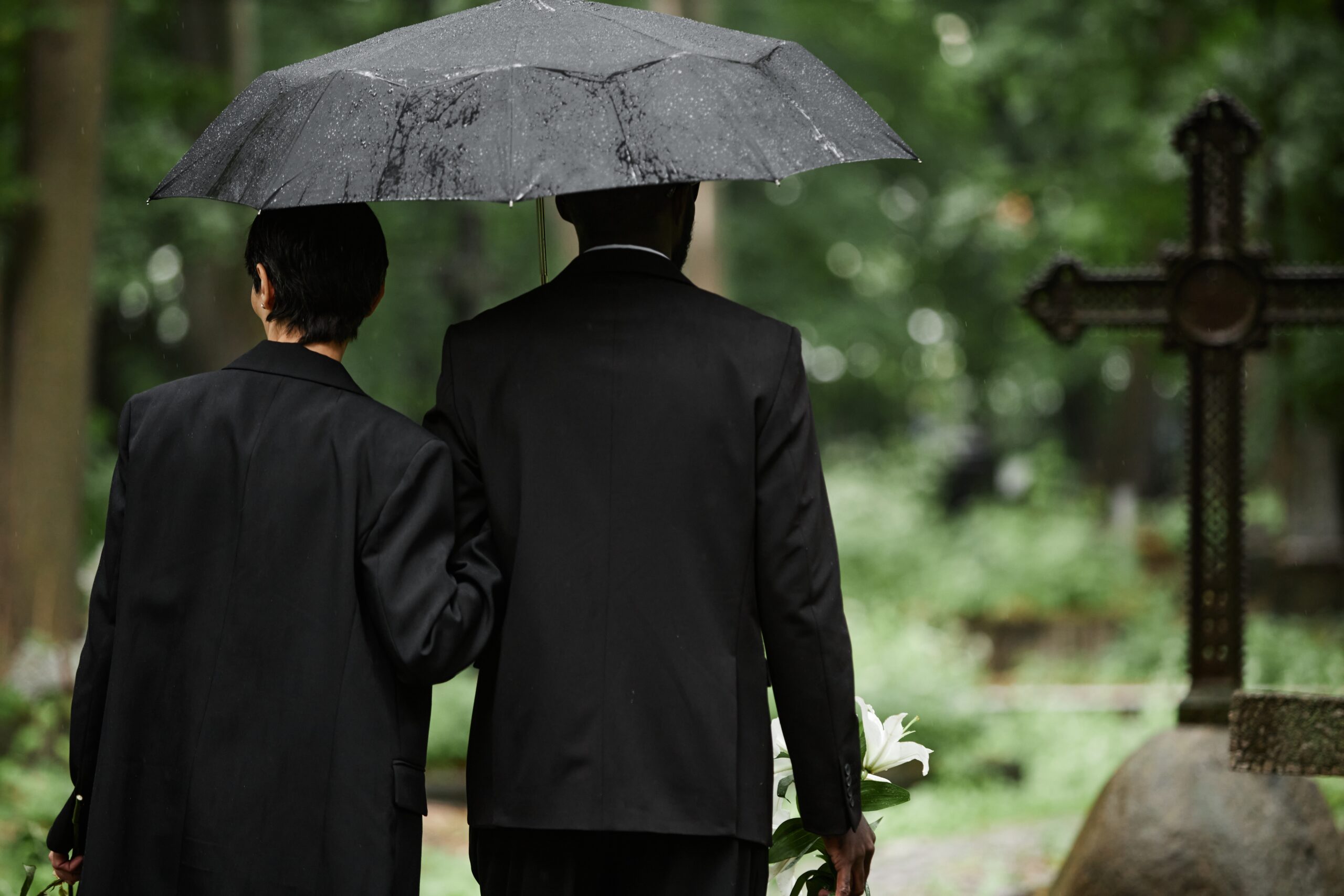 multiethnic couple in black suits walking under umbrella at cemetery
